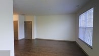 This single-floor space presents a straightforward, functional layout centered around an open living area. The room is finished with dark hardwood-style flooring that runs throughout, providing a warm contrast against the crisp white walls and bright white ceiling. The smooth, neutral wall surfaces offer a versatile canvas adaptable to a variety of interior styles and furnishing choices. Coordinating dark-toned baseboards and door trim add a cohesive design element that ties the room together. A paneled entry door in a dark finish is positioned along one wall, providing a defined point of entry. Multiple electrical outlets are conveniently distributed along the walls, supporting flexible furniture placement. The ceiling is clean and unadorned, contributing to the overall sense of openness in the room. The space is presented vacant, allowing for a clear assessment of its dimensions and layout. The overall ambiance is clean, modern, and neutral, making it well-suited for a range of uses.