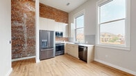Located at 1100 Washington Ave in Golden, Colorado, this thoughtfully designed 2-bedroom, 1-bathroom unit blends historic character with modern updates across a single floor. The kitchen is a standout feature, showcasing exposed brick walls that rise to tall ceilings, sleek cream cabinetry, stainless steel appliances including a French door refrigerator, a built-in microwave, a range, and a dishwasher. Light hardwood flooring and large windows allow natural light to pour in, offering views of the surrounding Golden streetscape. The hallway connects the living spaces efficiently and features stylish louvered and panel-style doors with matte black hardware, adding a contemporary design touch. Both bedrooms offer a calm, neutral palette — the primary bedroom features hardwood flooring and a ceiling fan, while the second bedroom is carpeted for added comfort. Each bedroom is served by its own dedicated closet. The full bathroom is sleekly appointed with a floating vanity, white countertop, tile flooring, and a glass-enclosed shower/tub combination. An in-unit laundry area adds everyday convenience. The exposed brick accent, visible across multiple rooms, ties the space together with a warm, industrial-chic aesthetic.