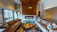 Located at 1600 Harbor Blvd in Weehawken, New Jersey, this sophisticated amenity lounge is designed with a contemporary aesthetic that blends warmth and modern elegance. The space is anchored by a striking linear gas fireplace set within a floor-to-ceiling warm wood panel surround, creating a dramatic focal point in the main seating area. Two plush leather sofas flank a natural wood slab coffee table, all resting atop a bold, abstract-patterned area rug that adds depth and visual interest to the space. Soaring ceilings amplify the sense of openness, while a sculptural multi-pendant chandelier provides ambient illumination above the lounge seating. Organic sculptural art pieces add a refined, curated touch throughout. A separate social area features a marble-topped bar counter with bar seating, accompanied by a dining arrangement with glass-top tables and mustard-toned mid-century modern chairs. Frosted glass sliding doors partition an additional private area, offering flexible use of the space. Large floor-to-ceiling glass doors and windows along one wall invite abundant natural light and open directly onto an outdoor terrace, seamlessly connecting the interior lounge to the exterior. The overall ambiance is polished yet inviting, ideal for gathering and relaxation.