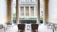 This remarkable space is defined by its exceptional neoclassical architecture, showcasing a covered colonnade terrace that bridges the elegance of historic design with modern comfort. Towering fluted Ionic columns frame the perimeter, creating a dramatic architectural backdrop that draws the eye upward along the soaring stone-clad walls. The flooring is laid in warm-toned natural stone tiles, lending a refined, cohesive aesthetic throughout.

Ornate arched niches punctuate the limestone walls, each adorned with classical pedestal urns filled with natural dried botanicals, reinforcing the space's sophisticated, timeless character. Generous seating arrangements are thoughtfully positioned throughout — woven-strap lounge chairs and plush grey upholstered sofas paired with minimalist black side tables create intimate conversation areas that contrast beautifully with the grand surroundings.

The colonnade opens outward to views of an adjacent neoclassical building facade featuring large multi-pane windows, decorative friezes, and further Ionic columns, extending the visual grandeur beyond the terrace itself. Lush green hedging and planted borders add a natural, softening element to the exterior boundary. The overall ambiance is one of quiet sophistication — a space that commands attention through architectural heritage while offering a welcoming, curated environment for relaxed gathering.