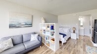 Located along Lankford Highway in Cape Charles, Virginia, this efficiently designed single-floor studio makes excellent use of its compact footprint. The open-plan layout seamlessly connects the living, sleeping, and kitchen areas, with light wood-look flooring running throughout to create a cohesive and airy feel.

The kitchen is well-equipped with white shaker-style cabinetry extending to the ceiling, granite-look countertops, a black electric range, an over-the-range microwave, and a stainless sink — offering all the essentials for comfortable everyday living. A textured ceiling adds subtle visual interest overhead.

The main living area is thoughtfully organized, with a open bookshelf unit serving as a stylish room divider that visually separates the sleeping area from the lounge space without closing off the room. A large picture window at the front of the unit allows abundant natural light to fill the space and provides an open view of the surrounding greenery outside.

The bathroom features a clean, white tub-shower combination with a decorative curtain, built-in soap and grab fixtures, and crisp white wall paneling, delivering a fresh and functional design.

This studio offers a bright, coastal-inspired atmosphere with a practical and efficient layout, ideally suited for those seeking a streamlined living space in the Cape Charles area.