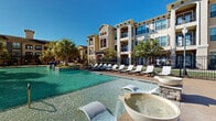 Located at 18545 University Blvd in Sugar Land, Texas, this outdoor amenity space showcases a resort-style design that combines Mediterranean-inspired architecture with thoughtfully planned recreational areas. The centerpiece is a generously sized swimming pool with crystal-clear turquoise water, complemented by distinctive water features including sculptural bowl fountains and cascading water elements that add both visual interest and a relaxing ambiance.

Surrounding the pool, a spacious sun deck is lined with contemporary white lounge chairs, providing ample space for relaxation under the Texas sun. Lush palm trees and mature landscaping frame the area, enhancing the resort atmosphere. A dedicated grilling and gathering zone features a paver-stone courtyard with covered pergolas supported by stone-clad columns and arched detailing, offering shaded outdoor dining and barbecue stations ideal for social gatherings.

The clubhouse building anchors the space with its arched entryways, lantern-style lighting, and warm stucco exterior — consistent with the community's cohesive Mediterranean aesthetic. Multi-story residential buildings with private balconies overlook the pool area, blending seamlessly into the overall design. The combination of water features, shaded retreats, and open lounging areas makes this outdoor space a well-appointed and inviting community amenity.