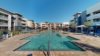 This outdoor amenity space is centered around a generously sized swimming pool with exceptionally clear, turquoise water, framed by a broad, stone-tiled sun deck. The pool area is flanked by multiple lounge chairs and blue market umbrellas, providing shaded relaxation spots along both sides of the water. A separate spa or wading area is integrated into the pool layout, offering a distinct space for leisure. The surrounding deck is finished in warm-toned, large-format stone pavers that extend throughout the entire outdoor area, lending a cohesive and upscale aesthetic.

The pool courtyard is enclosed by multi-story residential buildings featuring neutral stucco exteriors, private balconies, and large windows that overlook the amenity space. Mature trees with slender trunks are strategically placed throughout the deck, providing natural shade and softening the hardscape with greenery and low decorative plantings.

Additional seating nodes are distributed around the perimeter, including bar-height tables with chairs and intimate conversation sets shaded by striped blue umbrellas. A gated entrance area near the building provides a defined transition point, with outdoor dining furniture, ceiling fans, and manicured shrubs creating a welcoming atmosphere. The entire space benefits from abundant natural sunlight under open, clear skies.