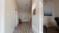 This well-appointed single-floor space combines cottage-inspired architectural details with a warm, functional layout. The entryway sets an inviting tone, featuring a wide hallway adorned with a decorative area rug, a classic chandelier overhead, and a painted shiplap ceiling that adds character and depth. Natural light pours in through a glass-paneled front door, illuminating the light-toned hardwood-style flooring that runs consistently throughout the space.

A console table with a mirror, decorative lanterns, and greenery lines one side of the hallway, offering a charming transitional zone between the entry and the main living areas. Multiple white paneled doors provide access to additional rooms, maintaining a clean and cohesive aesthetic.

The main gathering area features soaring vaulted ceilings with painted shiplap detailing and a ceiling fan, enhancing the airy feel of the room. A round dining table surrounded by upholstered chairs sits at the heart of this space, bathed in natural light from a side window and a set of elegant French doors that open to the outdoors. The French doors offer both a visual connection to the exterior and easy access to outside.

Overall, the space presents a cohesive, cottage-style aesthetic with warm neutral tones, thoughtful architectural features, and an abundance of natural light that creates a comfortable and welcoming atmosphere throughout.