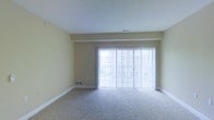 This single-floor space offers a clean, functional layout designed for comfortable living. The main living area is finished with wall-to-wall carpeting in a neutral tone, providing a soft and welcoming underfoot feel. Warm beige walls contribute to a calm, understated ambiance throughout the room.

A standout feature of the living area is the expansive sliding glass door framed by vertical blinds, which spans much of the exterior wall. This generous glazing floods the space with natural light and offers direct access to an outdoor area, creating a seamless connection between interior and exterior. The bright, airy quality of the room is further enhanced by the light-colored walls and ceiling.

A visible ledge or soffit detail along the upper portion of the wall adds a subtle architectural element to the space. Floor-level vents and white baseboards provide clean, practical finishing touches throughout. The overall design is straightforward and versatile, offering a neutral canvas well-suited to a variety of furnishing styles and personal preferences. The space presents a bright, open feel anchored by its prominent natural light source and simple, uncluttered layout.