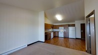 This single-floor space features an open-concept design that seamlessly connects the living area to the kitchen. The living area is finished with neutral-toned carpet, providing a comfortable and cohesive feel, while the kitchen transitions to durable hardwood-style flooring. The kitchen is well-appointed with warm wood-toned cabinetry offering generous upper and lower storage, white appliances including a refrigerator, electric range, and built-in microwave, as well as a sink set into a countertop along the perimeter. The L-shaped kitchen layout maximizes efficiency and workspace. Vaulted ceilings in the kitchen area add a sense of openness and volume to the space, enhanced by a flush-mounted ceiling light fixture that provides ample illumination. The transition between the living and kitchen areas is defined by a partial wall, creating a subtle separation while maintaining an airy, open feel. A baseboard heater runs along the living area wall, indicating a functional heating system. The overall aesthetic is practical and clean, with a neutral color palette throughout.