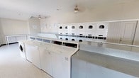 Located at 3226 Persimmon Rd in Dallas, TX, this single-story commercial laundry facility is designed for efficient, high-volume use. The main laundry area is generously proportioned, housing a row of top-load washing machines positioned along a central island with a durable speckled laminate folding counter. Along the back wall, multiple stacked dryer units are arranged in a uniform configuration, providing substantial drying capacity. Ceiling fans are installed throughout the laundry room to maintain air circulation, and recessed vents support additional ventilation. Instructional signage is posted on the walls for patron guidance.

The flooring throughout the facility consists of durable, easy-to-clean vinyl tile, practical for a high-traffic environment. A separate corridor area connects to the laundry room, featuring paneled walls painted in a neutral gray tone, a built-in bench for seating, and large windows that allow natural light to filter in and offer views of the surrounding outdoor area. The hallway also includes additional counter space and leads to a secondary room, providing further functional utility. Fluorescent lighting ensures consistent illumination throughout all areas. The overall layout is clean, organized, and purpose-built for commercial laundry operations.
