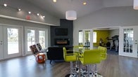 This single-floor space showcases a thoughtfully designed open-concept layout that blends comfort and modern style. The main lounge area is anchored by a sleek wall-mounted electric fireplace set against a bold dark accent wall, complemented by a wall-mounted flat-screen TV. The space is furnished with a contemporary black sofa, checkered accent chairs, and a striking orange sculptural side table, creating an inviting and eclectic atmosphere.

A high-top bar-style table surrounded by vibrant chartreuse green swivel chairs adds a lively social element to the room. The warm hardwood-style flooring ties the space together, while a large pendant drum light fixture provides a focal point overhead. Track lighting further enhances the ambiance throughout.

Natural light floods the room through large glass panel doors along one wall, offering views of an outdoor area and lending an airy, open feel to the interior. A large mirror positioned near the entry to an adjoining room expands the sense of space visually.

Adjacent to the main lounge, a fitness room is visible through mirrored double doors, featuring cardio equipment set against a bright yellow-green accent wall. The overall design palette — charcoal, gray, and bold lime green — creates a dynamic, energetic environment ideal for both relaxing and socializing.