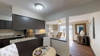 This single-story residence offers a well-organized layout that connects key living areas in a practical and cohesive flow. The kitchen is outfitted with dark cabinetry, granite-style countertops, tile flooring, and stainless steel appliances including a refrigerator, dishwasher, and electric range. A pass-through counter opens directly to a cozy dining nook, where a small round table sits beneath a flush-mount ceiling light, creating an intimate space for casual meals.

Adjacent to the kitchen, the living room is the most expansive area of the unit, featuring rich hardwood-style flooring, a ceiling fan with a light fixture, and a sliding glass door that allows natural light to fill the space. The room accommodates a comfortable seating arrangement and is accented by large framed artwork along the walls.

The bedroom is carpeted and well-proportioned, offering space for a full-sized bed and nightstands, with a window providing natural light and a closet nearby for storage. The bathroom includes a tub and shower combination with neutral tile surround, a decorative mosaic accent border, tile flooring, and a granite-topped vanity, presenting a clean and updated appearance throughout.
