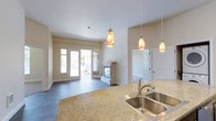 This single-floor space offers an open-concept layout that seamlessly connects the kitchen and living areas, creating a welcoming and functional environment. The kitchen features granite countertops with warm gold and beige tones, a stainless steel sink, and a series of decorative pendant lights with amber-toned glass shades that cast a warm glow over the workspace. These pendants serve as a visual anchor, tying the kitchen and living space together cohesively.

The living area is highlighted by a stylish fireplace with a clean white mantel, providing a focal point that adds both warmth and character to the room. Large French doors flood the space with natural light and offer direct access to an outdoor area, enhancing the sense of openness and connectivity to the exterior. The neutral color palette of soft whites and warm beiges throughout the walls contributes to an airy, inviting atmosphere.

Durable wood-look flooring extends across the living area, offering a practical yet attractive finish. A separate door visible from the main living space suggests additional rooms or storage. Overall, the layout is thoughtfully designed to maximize both comfort and flow.