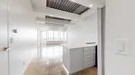 This contemporary 3-bedroom, 2-bathroom residence showcases an industrial-modern aesthetic defined by polished concrete floors throughout, crisp white walls, and architecturally expressive exposed ceiling elements including metal decking and structural beams. The open-concept living area flows seamlessly into the kitchen, which features a gray island with a white countertop, white cabinetry, a subway tile backsplash, and stainless steel appliances — including a refrigerator with an ice and water dispenser. Recessed lighting illuminates the shared spaces, complementing the abundant natural light that pours in through large windows framing impressive urban skyline views. The generously sized primary bedroom offers polished floors, mirrored closet doors, and direct access to natural light and city views. A dedicated walk-in closet provides wire-rack shelving for ample storage. The two bathrooms continue the clean, modern palette with gray vanities and white finishes. Additional bedrooms are well-proportioned with recessed lighting and polished concrete floors. Throughout the residence, the interplay of white surfaces, industrial ceiling details, and reflective flooring creates a bright, cohesive, and distinctly urban living environment.