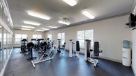 The fitness center is a bright, single-story space designed to support a well-rounded workout routine. The interior features white walls and a neutral, dark-toned rubberized floor, creating a clean and functional environment suited for exercise. Ceiling fans are mounted overhead alongside flush rectangular light fixtures, ensuring the space remains well-ventilated and well-lit throughout the day.

Large windows line the walls, allowing natural light to pour in and offering views of surrounding greenery, contributing to an open and refreshing atmosphere. The equipment is thoughtfully arranged to maximize usable floor space while maintaining comfortable circulation between machines.

The fitness center includes a selection of cardio equipment such as stationary bikes and elliptical trainers, as well as cable-based strength-training machines designed for upper and lower body workouts. An adjustable bench is also present, adding versatility for a range of exercises. The combination of white and black equipment gives the space a cohesive, modern aesthetic. Overall, the fitness center provides a clean, accessible, and adequately equipped environment for residents or members seeking a convenient on-site workout facility.