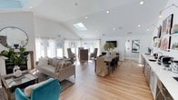 This single-floor space showcases an open-concept layout that seamlessly integrates a living area and dining space into one cohesive, light-filled environment. The vaulted ceiling rises to an impressive peak, amplifying the sense of spaciousness, while a centrally positioned skylight floods the interior with abundant natural light throughout the day. Recessed lighting complements the natural illumination, ensuring the space remains bright at all hours.

The flooring throughout is light-toned hardwood, contributing to the clean, contemporary aesthetic. The living area is anchored by a neutral-toned sofa paired with a bold teal accent chair, adding a pop of color against the predominantly white and beige palette. A chevron-patterned area rug grounds the seating arrangement.

The dining area features a long wooden table surrounded by a mix of slipcovered and upholstered chairs in complementary neutral tones. A decorative vase with greenery serves as a natural centerpiece. Large French doors and floor-to-ceiling windows line the far wall, offering generous outdoor views and enhancing the airy, welcoming ambiance of the space.