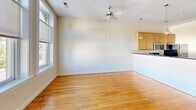 Located at 209 E Main St in the heart of downtown Richmond, Virginia, this single-floor unit offers a clean, open-concept layout that seamlessly connects the living area and kitchen. Warm hardwood flooring flows consistently throughout the space, lending a cohesive and inviting feel to every room.

The kitchen is a standout feature, outfitted with light maple cabinetry, dark granite countertops, and a full suite of stainless steel appliances including a refrigerator, electric range, and over-the-range microwave. A peninsula-style breakfast bar with a low half-wall divider defines the kitchen space while maintaining an open sightline to the living area. A series of pendant lights hang above, providing both focused task lighting and a modern aesthetic touch.

The living area is generously proportioned, featuring a ceiling fan and large windows that allow natural light to fill the room, offering views of the surrounding urban streetscape. The entryway is framed by an arched interior door and accompanied by an additional window that further brightens the space.

A separate room with darker hardwood flooring provides a quieter, more enclosed area that can function as a bedroom or private retreat. Throughout the unit, neutral white walls and clean architectural lines create a versatile backdrop suited to a variety of uses in the heart of Richmond's downtown corridor.