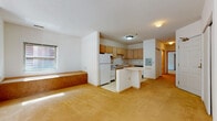 This single-floor unit is designed with an open, practical layout that connects the living area and kitchen in one continuous space. The living area is finished with warm, neutral carpeting and features a built-in bench along the exterior wall beneath a window, which allows natural light to fill the room. A textured ceiling runs throughout the main living space, contributing to a clean, understated aesthetic.

The kitchen is positioned centrally within the open floor plan and is equipped with light wood cabinetry offering generous storage, a white refrigerator, a gas range with overhead ventilation, a sink, and a black dishwasher. Light-toned tile flooring defines the kitchen zone. A peninsula with a curved countertop edge extends into the living space, creating a natural division while maintaining visual openness. An electrical panel is mounted on the kitchen wall for convenient access.

A hallway leads to the remaining rooms, including a carpeted bedroom that receives natural light through a window and offers a quiet, simple space. The bathroom is outfitted with accessibility features, including stainless steel grab bars near the toilet and a roll-under vanity. In-unit laundry is conveniently located within the bathroom, adding everyday practicality to the space.
