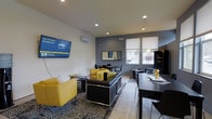 This single-floor commercial space is designed with a clean, contemporary aesthetic that balances professionalism with comfort. The main waiting and reception area serves as the focal point of the space, featuring a striking color palette of bold yellow and black that gives the interior a vibrant, energetic feel.

The seating area is furnished with plush yellow accent chairs and a sleek black leather sofa, arranged around a patterned area rug that adds visual depth to the light hardwood flooring beneath. A wall-mounted flat-screen television displaying branded content anchors one wall, making it an ideal feature for client engagement while they wait.

A wall-mounted mini-split HVAC unit ensures year-round climate comfort, while recessed ceiling lighting provides consistent, even illumination throughout. A two-tone wall design—crisp white above and a deep charcoal-gray accent below—adds architectural character and a modern edge to the interior.

Natural light flows in through roller-shade windows along one wall, offering views of the exterior surroundings. A dark wood dining or consultation table with bar-height seating is positioned near the windows, providing a functional space for meetings or informal gatherings. A coordinating dark wood shelving unit and side tables complete the refined, cohesive look of this well-appointed professional environment.