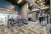 This fitness center combines industrial-modern aesthetics with a comprehensive range of workout equipment, creating a functional and visually dynamic environment. The space is defined by its exposed concrete columns, bold teal accent walls, and large windows that invite natural light and outdoor views, contributing to an energizing atmosphere throughout.

The flooring consists of herringbone-patterned carpet tiles in neutral tones of beige, gray, and blue, adding visual texture underfoot. Overhead, exposed large-diameter ductwork and track lighting run across the dark ceiling, reinforcing the industrial character of the space.

The cardio zone features a row of treadmills and elliptical machines positioned along the windowed perimeter, allowing users to exercise with an outward view. A dedicated strength training area houses squat racks, weight benches, and plate-loaded barbells, while a selection of selectorized machines and a tiered dumbbell rack serve the free weights section. A punching bag and additional functional training accessories round out the equipment offerings.

A separate glass-enclosed room provides a quieter workout zone, and a vending station with beverages and snacks adds convenience. Wall-mounted televisions are positioned throughout for entertainment.