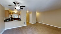 This two-floor residential space is designed with practicality and comfort in mind. The main living area is generously proportioned, featuring warm beige carpeting that runs throughout, creating a cohesive and inviting atmosphere. Neutral warm-toned walls provide a versatile backdrop, complemented by clean white baseboards and trim.

A notable feature of the living area is the ceiling fan with an integrated light fixture, which provides both air circulation and ambient lighting. The space is further illuminated by wall-mounted light fixtures positioned at key points throughout the room, ensuring consistent brightness.

An open pass-through counter connects the living area to the kitchen, where a dark granite or stone countertop is visible alongside cabinetry and a window that allows natural light to filter in. This open-concept connection between the kitchen and living space promotes an easy flow for everyday living and entertaining.

A hallway accessible from the main living area leads to additional rooms on the same floor, while a second floor offers further living space. The layout includes built-in wall vents for climate control, reinforcing the home's functional design. Overall, this space presents a straightforward, well-maintained residential environment well-suited for comfortable everyday living.