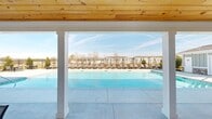 Located at 720 Curtis Pkwy SE in Calhoun, Georgia, this three-floor community offers a well-appointed outdoor amenity area designed for both relaxation and social gathering. The centerpiece of the outdoor space is a generously sized swimming pool with crystal-clear blue water, framed by a broad concrete pool deck lined with rows of cushioned lounge chairs — providing ample space for sunbathing and outdoor leisure.

A covered cabana structure anchors one side of the pool area, featuring clean white columns and a warm natural wood-paneled ceiling that adds a refined, welcoming character. The covered space offers a shaded retreat directly adjacent to the water. On the opposite end, a clubhouse-style building with board-and-batten siding, a covered porch, and large windows overlooks the pool, providing additional gathering and indoor-outdoor connectivity.

An open patio area adjacent to the pool features multiple seating arrangements with round tables and chairs, enclosed by a sleek black iron fence that defines the perimeter while preserving open views of the surrounding landscape and the multi-story residential buildings beyond. The overall outdoor environment is clean, modern, and spacious, reflecting a community-oriented design suited to the Calhoun, Georgia setting.