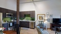 Located at 300 Blackwell Street in Durham, North Carolina, this single-floor space offers a compelling blend of historic industrial character and contemporary design. The open-concept layout is anchored by a striking exposed timber column, a nod to the building's storied past, while soaring ceilings lend an expansive, airy feel throughout.

The kitchen area is thoughtfully appointed with dark espresso cabinetry, a bold green glass tile backsplash, stainless steel appliances, and light-toned countertops. The cabinetry extends to the ceiling, maximizing storage while maintaining a clean, modern aesthetic. A stainless steel dishwasher and gas range complete the functional kitchen setup.

Light hardwood flooring runs throughout the space, warming the industrial aesthetic and creating a cohesive visual flow between the kitchen and the open living area. The living space is versatile and well-lit, with high white walls that serve as a natural gallery for artwork, as seen with the gold-framed painting displayed near a classic writing desk and accent chair.

Track lighting overhead allows for flexible illumination across the space. The combination of raw architectural elements, such as the timber post and tall ceilings, with refined modern finishes creates a distinctive, loft-inspired ambiance well-suited to a variety of uses.
