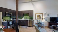 Located at 300 Blackwell Street in Durham, North Carolina, this single-floor space offers a compelling blend of historic industrial character and contemporary design. The open-concept layout is anchored by a striking exposed timber column, a nod to the building's storied past, while soaring ceilings lend an expansive, airy feel throughout.

The kitchen area is thoughtfully appointed with dark espresso cabinetry, a bold green glass tile backsplash, stainless steel appliances, and light-toned countertops. The cabinetry extends to the ceiling, maximizing storage while maintaining a clean, modern aesthetic. A stainless steel dishwasher and gas range complete the functional kitchen setup.

Light hardwood flooring runs throughout the space, warming the industrial aesthetic and creating a cohesive visual flow between the kitchen and the open living area. The living space is versatile and well-lit, with high white walls that serve as a natural gallery for artwork, as seen with the gold-framed painting displayed near a classic writing desk and accent chair.

Track lighting overhead allows for flexible illumination across the space. The combination of raw architectural elements, such as the timber post and tall ceilings, with refined modern finishes creates a distinctive, loft-inspired ambiance well-suited to a variety of uses.