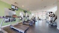 This single-floor fitness center is designed to provide a comprehensive workout environment, combining functionality with a bright, welcoming atmosphere. The space is organized to accommodate a variety of exercise modalities, with distinct zones for cardio training, strength equipment, and open-floor stretching or functional fitness activities.

The cardio area features multiple treadmills and elliptical machines from SportsArt, positioned along green accent walls accented by large mirrors that visually expand the space. Wall-mounted flat-screen televisions are positioned above the cardio equipment, allowing users to stay entertained during workouts. Recessed lighting throughout the ceiling supplements the abundant natural light that pours in through the expansive floor-to-ceiling windows along the front of the facility, offering views of the outdoor surroundings.

The strength training section includes a multi-functional cable machine, a weight bench, and a stability ball, along with additional resistance equipment. Rubber flooring spans the entire space, providing both durability and safety underfoot. A water dispenser is conveniently stationed near the windowed facade. The clean white walls, high ceilings, and generous glazing create an open, airy ambiance throughout the fitness center.