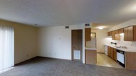 This single-floor residential space offers a straightforward and functional layout designed for comfortable everyday living. The living area is generously sized, featuring wall-to-wall carpeting in a neutral tone that adds warmth underfoot. A textured popcorn ceiling and cream-colored walls create a clean, understated backdrop throughout. HVAC vents are integrated into both the ceiling and lower walls, ensuring efficient climate control across the space. A solid wood-paneled interior door provides access to an additional room, adding to the unit's practical layout. Transitioning from the living area, the kitchen is open and connected, defined by classic wood-toned cabinetry that offers ample storage. Light countertops provide a contrasting work surface, and a white refrigerator and additional appliances complete the space. The kitchen features hard flooring, offering a practical and easy-to-maintain surface distinct from the carpeted living area. Overhead lighting in the kitchen ensures a well-lit environment for meal preparation. Overall, the space presents a no-frills, utilitarian design with all the essential elements needed for comfortable day-to-day living.