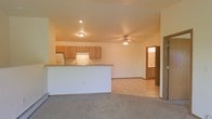 This single-floor space is designed with an open-concept layout that seamlessly connects the kitchen and living area, creating a functional and cohesive environment. The kitchen is equipped with medium-toned wood cabinetry, a white electric range with an overhead ventilation hood, and a refrigerator. A peninsula counter with a raised breakfast bar extends into the living space, offering additional workspace and casual seating while visually defining the two areas.

The living area features neutral-toned carpeting and smooth, light-colored walls that contribute to a clean, airy atmosphere. A ceiling fan with a light fixture is centrally positioned overhead, providing both air circulation and ambient lighting. Recessed lighting in the kitchen area adds focused illumination. The flooring transitions from carpet in the living space to a hard surface in the kitchen area, providing practical zoning between the two spaces.

Visible through a doorway at the far end of the living area is a bathroom, outfitted with warm wood-toned cabinetry and a vanity. The overall color palette throughout the space is neutral and understated, with beige and cream tones dominating the walls, ceilings, and floors, lending the interior a simple, practical character.