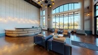 Located on Herb Hill Road in Glen Cove, NY, this elegantly designed lobby makes a striking first impression with its soaring double-height ceilings and floor-to-ceiling windows that flood the space with natural light and offer expansive outdoor views. At the heart of the entry is a grand curved reception desk, set against a dramatic floor-to-ceiling stone-clad accent wall with cascading pendant lights that create a warm, inviting atmosphere.

The main lobby area features multiple seating arrangements with deep blue upholstered sofas and armchairs paired with a live-edge wood coffee table, all anchored by an ornate chandelier overhead. A decorative laser-cut wall panel adds an artistic architectural touch near the entrance.

Adjacent to the main reception area, a more intimate lounge zone offers diverse seating options, including a high-backed upholstered banquette set against a rich wood slat feature wall, along with sleek armchairs arranged around small accent tables. This area flows into a cozy sitting room appointed with vibrant yellow accent chairs, a linear electric fireplace set into a textured stone wall, and a built-in shelving unit housing a flat-screen TV and decorative objects.

A compact coffee bar area completes the space, outfitted with a professional espresso machine, counter seating at a butcher-block island, and a large window overlooking the outdoors — ideal for casual gatherings.
