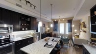 This single-floor space showcases a thoughtfully designed open-concept layout that seamlessly connects the kitchen, dining, and living areas into one cohesive environment. The kitchen is a standout feature, equipped with dark espresso cabinetry, a light granite-topped island with bar seating, a subway tile backsplash, and stainless steel appliances. Pendant lighting hangs above the island, complemented by track lighting overhead, creating a well-lit and functional workspace. Open shelving on the upper cabinetry adds a decorative touch, displaying curated décor accents.

The flow from the kitchen leads naturally into the dining and living areas, where hardwood-style flooring runs throughout, tying the spaces together. The living area benefits from large French doors that invite natural light deep into the interior while providing access to the outdoors. Sheer and drape curtain combinations frame the doors elegantly. A defined dining space sits between the kitchen and living room, offering flexibility for both casual and formal use. The overall ambiance is modern and refined, with a warm, inviting atmosphere created by layered lighting and cohesive, contemporary finishes throughout.