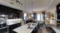 This single-floor space showcases a thoughtfully designed open-concept layout that seamlessly connects the kitchen, dining, and living areas into one cohesive environment. The kitchen is a standout feature, equipped with dark espresso cabinetry, a light granite-topped island with bar seating, a subway tile backsplash, and stainless steel appliances. Pendant lighting hangs above the island, complemented by track lighting overhead, creating a well-lit and functional workspace. Open shelving on the upper cabinetry adds a decorative touch, displaying curated décor accents.

The flow from the kitchen leads naturally into the dining and living areas, where hardwood-style flooring runs throughout, tying the spaces together. The living area benefits from large French doors that invite natural light deep into the interior while providing access to the outdoors. Sheer and drape curtain combinations frame the doors elegantly. A defined dining space sits between the kitchen and living room, offering flexibility for both casual and formal use. The overall ambiance is modern and refined, with a warm, inviting atmosphere created by layered lighting and cohesive, contemporary finishes throughout.