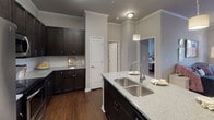 This single-floor residence offers a thoughtfully designed layout with a cohesive aesthetic defined by neutral gray walls, dark espresso cabinetry, and light granite-style countertops throughout. The kitchen serves as a functional centerpiece, featuring dark shaker-style upper and lower cabinets extending nearly to the ceiling, stainless steel appliances including a refrigerator, range, and over-the-range microwave, and a white subway tile backsplash. A large island with a double undermount sink and pendant lighting adds both workspace and visual appeal, complemented by rich hardwood flooring and crown molding details.

The living area provides a comfortable, well-lit space with natural light streaming through large windows dressed with blinds and curtains. Crown molding adds a refined touch to the room. Multiple bedrooms feature plush carpeting, ceiling fans, and windows with outdoor views, creating bright and restful retreats. The primary bedroom includes multiple doors suggesting access to a private bath or walk-in closet.

The bathrooms are consistently finished with the same dark vanity cabinetry, granite-topped counters, tile flooring, and a shower-tub combination with a curved curtain rod, maintaining the home's unified design language throughout.