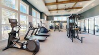 Located at 9500 Grandtree Ave in Venice, Florida, this fitness center offers a generously sized, single-floor layout designed to support a comprehensive workout experience. The space is divided into two distinct zones, seamlessly connected through a glass-panel partition wall that adds a modern, open feel while providing visual separation between areas.

The main workout area is defined by its striking architectural design, featuring elevated tray ceilings accented with warm, exposed wood beams and an oversized ceiling fan. Recessed lighting throughout ensures the space is well-lit at all times. The walls are finished in a bold deep teal above white wainscoting, creating a clean and polished aesthetic. Large windows line the perimeter, flooding the floor with abundant natural light.

The cardio zone is equipped with a row of treadmills, elliptical machines, a stair climber, and stationary spin bikes. The free weights and strength training section features a full power rack with weight plates, a dumbbell rack, and a multi-function cable machine. A dedicated spin and stretching room offers floor-to-ceiling windows framing tranquil water and natural landscape views, creating a serene backdrop for cycling and low-impact training. Light-toned carpet flooring runs throughout, adding comfort underfoot across the entire facility.