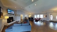 This well-appointed single-floor space offers an open, airy layout highlighted by vaulted ceilings with crown molding and rich, dark hardwood flooring throughout. The main living area is generously sized, centered around a cozy fireplace and furnished with a large blue sectional sofa paired with a patterned area rug, creating a comfortable gathering zone. Recessed lighting and a ceiling fan provide both practical illumination and climate comfort overhead, while a textured ceiling adds architectural character.

Toward the rear of the space, a dedicated seating lounge features leather armchairs arranged over a geometric rug, positioned near a wall of large windows that allow ample natural light to pour in and offer views to the outdoors. Adjacent to this lounge sits a functional office nook outfitted with a desk, task chair, and a floor lamp, making the space versatile for both relaxation and productivity.

The soft blue-grey walls create a calm, neutral backdrop that complements the warm tones of the hardwood floors and furnishings. An emergency exit door and wall-mounted television are also present, suggesting this space serves as a shared or communal facility. Overall, the design balances modern comfort with practical functionality in a bright, welcoming environment.