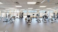 Located at 1121 W Prospect Rd in Fort Collins, Colorado, this well-appointed fitness facility offers a single-floor exercise room designed to support a wide range of workout needs. The space features a drop-panel ceiling with recessed fluorescent lighting and ceiling fans that maintain a bright, comfortable environment throughout.

The cardio section is generously equipped with multiple treadmills, elliptical trainers, stair climbers, and spinning bikes, all arranged to allow ample space between machines. Several flat-screen televisions are mounted along the walls, providing entertainment during workouts. A large mirror runs along one wall, enhancing the sense of openness in the space.

The strength training area includes a multi-station cable machine, a squat/power rack, adjustable benches, and a full dumbbell rack ranging across various weights, providing options for both beginner and advanced lifters. The durable rubber flooring throughout the facility ensures safety and sound absorption.

Large windows along the perimeter allow natural light to filter into the space and provide views of the surrounding outdoor environment. The overall layout is thoughtfully organized, with clear zones for cardio and strength training that allow multiple users to exercise simultaneously without congestion. The clean, neutral color palette and practical design create a focused and functional fitness environment.