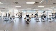 Located at 1121 W Prospect Rd in Fort Collins, Colorado, this well-appointed fitness facility offers a single-floor exercise room designed to support a wide range of workout needs. The space features a drop-panel ceiling with recessed fluorescent lighting and ceiling fans that maintain a bright, comfortable environment throughout.

The cardio section is generously equipped with multiple treadmills, elliptical trainers, stair climbers, and spinning bikes, all arranged to allow ample space between machines. Several flat-screen televisions are mounted along the walls, providing entertainment during workouts. A large mirror runs along one wall, enhancing the sense of openness in the space.

The strength training area includes a multi-station cable machine, a squat/power rack, adjustable benches, and a full dumbbell rack ranging across various weights, providing options for both beginner and advanced lifters. The durable rubber flooring throughout the facility ensures safety and sound absorption.

Large windows along the perimeter allow natural light to filter into the space and provide views of the surrounding outdoor environment. The overall layout is thoughtfully organized, with clear zones for cardio and strength training that allow multiple users to exercise simultaneously without congestion. The clean, neutral color palette and practical design create a focused and functional fitness environment.