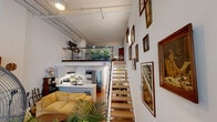 Located in Vancouver's vibrant Mount Pleasant neighbourhood, this two-level loft offers a thoughtfully designed layout across two distinct floors. The main level houses an open-concept kitchen and living area bathed in natural light from large street-facing windows. The kitchen features white cabinetry, a stainless steel refrigerator, and a peninsula with a sink, flowing seamlessly into the living space below soaring ceilings with exposed industrial pipework and track lighting. A striking open-tread staircase with a glass-panel railing connects the two levels, adding a contemporary architectural focal point. The main floor also includes a dedicated laundry area and an in-unit bathroom complete with a bathtub and shower. Ascending to the upper level, the spacious bedroom accommodates a full sleeping and workspace, with a mirrored closet adding depth to the room. Exposed ductwork and continued track lighting maintain the industrial aesthetic throughout. A private patio extends the living space outdoors, offering a welcome retreat. The loft's design balances urban industrial character with functional, comfortable living.