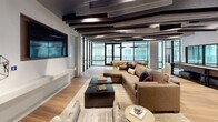 This single-floor amenity space is defined by its contemporary design and thoughtfully curated entertainment zones. The lounge area serves as the centerpiece, furnished with a generously sized sectional sofa, accent pillows, and a dark coffee table set atop a neutral area rug. A wall-mounted flat-screen television anchors one end of the room, while a sculptural ceiling installation of layered acoustic panels in dark tones adds dramatic visual interest overhead. Warm wood-look flooring runs throughout, providing a cohesive foundation across all areas.

The game room is well-appointed, featuring a full-size billiards table positioned beneath pendant lighting and a dedicated shuffleboard table along a mirrored wall. A wet bar with dark stone countertops, wood cabinetry, and a stainless steel refrigerator serves the entertainment space with convenience.

Expansive floor-to-ceiling glass walls and black-framed curtain wall systems flood the interior with natural light while offering panoramic views of the surrounding courtyard and pool area below. The outdoor balcony access further enhances the open, connected ambiance of this stylish communal retreat.
