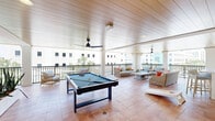 Located at 125 Quadrille Plaza Dr in West Palm Beach, FL, this covered rooftop amenity terrace offers a well-appointed social and recreation space designed for both relaxation and entertainment. The terrace spans a generous single-floor footprint, finished throughout with warm terracotta tile flooring that complements the bright, coastal aesthetic.

A striking wood-paneled ceiling runs the length of the space, lending warmth and architectural character overhead, while multiple ceiling fans ensure comfortable airflow in the Florida climate. Sturdy white columns frame open balcony edges lined with decorative wrought-iron railings, providing sweeping views of the West Palm Beach skyline and surrounding urban landscape.

The recreation zone is anchored by a full-size pool table and is complemented by a cornhole set, catering to a range of social activities. A generously sized lounge area features wicker and upholstered seating with vibrant accent pillows, arranged around low coffee tables for easy conversation. Lush snake plant arrangements in raised white planters add greenery and a clean, modern touch throughout.

An outdoor kitchen and bar area, accented with a decorative mosaic tile feature wall, provides a convenient space for entertaining. High-top bistro tables nearby offer additional casual seating. The overall ambiance is light, airy, and resort-inspired, making this terrace an ideal gathering venue.