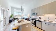 Located at 2132 City Gate Ln in Naperville, IL, this contemporary single-floor residence combines modern design with functional living. The open-concept layout seamlessly connects the kitchen, dining, and living areas beneath impressively soaring ceilings that amplify the sense of space.

The kitchen is outfitted with sleek gray flat-front cabinetry extending to ceiling height, stainless steel appliances including a gas range, built-in microwave, dishwasher, and refrigerator, all complemented by crisp white quartz countertops. A central island with bar seating provides additional prep space and casual dining. Striking linear pendant lighting anchors the kitchen and adds a contemporary design touch.

The living area is bright and airy, featuring large floor-to-ceiling windows that flood the space with natural light and offer open outdoor views. Light-toned hardwood flooring runs throughout the main living areas, enhancing the clean, modern aesthetic.

The bedroom is a calm retreat highlighted by a bold teal accent wall, plush carpeting, large windows, and ample natural light. A second bedroom offers a similarly serene atmosphere with neutral tones and outdoor views.

The bathroom features full-height tile surrounding a soaking tub and shower combination, a clean white vanity with an under-mount sink, and wood-toned flooring, delivering a polished, spa-like finish.