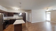 This single-floor space showcases an open-concept layout that seamlessly connects the kitchen and living areas, creating a cohesive and functional environment. The kitchen is a standout feature, equipped with rich dark espresso cabinetry that extends to include a distinctive diamond-pattern wine rack for added character. The countertops are finished in light granite, offering a striking contrast to the dark cabinetry. A central kitchen island with a matching granite surface provides additional workspace and serves as a natural dividing point between the kitchen and the adjoining living area. Pendant lighting hangs above the island, complementing the track lighting positioned over the kitchen workspace to ensure well-lit food preparation areas. Stainless steel appliances complete the kitchen's modern aesthetic. The flooring throughout is a warm-toned hardwood, lending continuity and a sense of warmth across the entire open space. The living area is generously sized with neutral-toned walls, offering a versatile blank canvas for various furniture arrangements. A closeted door leads to an additional room or utility space, adding to the unit's practical functionality.
