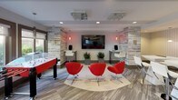 Located at 11351 Rockville Pike in Rockville, Maryland, this single-floor amenity space offers a well-designed, modern environment with distinct functional zones. The entrance features floor-to-ceiling glass doors framed in dark metal, flooding the entry corridor with natural light and providing views of an outdoor courtyard with mature landscaping. Light wood-look flooring and clean white walls establish a cohesive, contemporary aesthetic throughout.

The co-working and social lounge area centers around a large, white quartz-topped island bar with dark cabinetry, surrounded by modern bar seating. Sculptural pendant lights with honeycomb shades hang overhead, while a wall-mounted television and built-in kitchenette with a stainless steel sink, cabinetry, and under-counter appliances provide convenience. Colorful map-inspired artwork adds personality to the space.

A formal conference area features a striking elongated white table with wood inlay detailing, flanked by seating for multiple occupants, a wall-mounted display screen, bold red abstract artwork, and a dedicated workstation along the perimeter.

The entertainment lounge contrasts with reclaimed wood-style accent walls, a large flat-screen television, sound system, crystal ceiling fixtures, and vibrant red swivel chairs arranged around a central rug. A foosball table adds a recreational element, completing the dynamic and versatile character of the space.