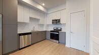 This single-floor unit offers a clean, modern aesthetic throughout, with light wood-tone flooring and crisp white walls that create a bright, cohesive atmosphere across all spaces.

The kitchen is a standout feature, designed with a practical L-shaped layout. Two-toned cabinetry — white upper cabinets and grey lower cabinets — is paired with a classic white subway tile backsplash that extends across the entire cooking area. Stainless steel appliances include a gas range, an over-the-range microwave, and a dishwasher. Light stone countertops and a single-basin sink with a modern faucet complete the functional workspace.

The bathroom maintains the same contemporary style, featuring a white vanity with a marble-patterned stone countertop and an integrated sink. A large mirror above the vanity enhances the sense of space, while the light wood flooring continues seamlessly from the main living areas. The bathroom also provides access to a dedicated closet area with built-in shelving, offering organized storage.

The bedroom is a calm, minimalist space with neutral white walls and light wood flooring. A window with horizontal blinds allows for natural light while maintaining privacy. The unit's overall design prioritizes clean lines, neutral tones, and practical finishes, making it a well-appointed and move-in ready space.