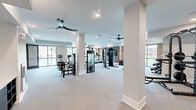 Located at 501 W Waugh St in Dalton, Georgia, this well-appointed fitness center offers a spacious, single-floor layout designed to support a full range of workout activities. The open floor plan is anchored by clean white walls, light-toned flooring, and structural columns that define distinct exercise zones without disrupting the sense of openness. Recessed lighting throughout, paired with multiple ceiling fans, ensures the space remains bright and comfortable during any workout.

The cardio area is equipped with treadmills, ellipticals, and stationary bikes positioned along large floor-to-ceiling windows that frame views of the surrounding streetscape, flooding the zone with natural light. A dedicated strength training section features a multi-station cable machine, a squat rack with barbell and weight plates, adjustable benches, and a full dumbbell rack with neatly organized free weights.

A distinctive accent wall featuring a herringbone-patterned tile design and warm wood-paneled sections adds a modern aesthetic touch to the interior. A water station and storage shelving are conveniently positioned near the entrance. The space also includes a mirrored wall to assist with form and technique. Double glass doors provide easy access, completing a fitness facility that balances functionality with contemporary design.