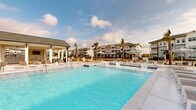 Located on Browning Way in Foley, Alabama, this outdoor amenity space delivers a resort-style experience designed for relaxation and recreation. The centerpiece is a generously sized swimming pool with crystal-clear blue water, featuring a gradual sun shelf entry that allows residents to lounge partially submerged in the shallow water. The pool deck is finished in a light-toned concrete surface and lined with an extensive collection of modern white-framed chaise lounges, providing ample space for sunbathing. Striped black-and-white market umbrellas are positioned throughout the deck to offer shaded retreats. A well-appointed single-story cabana structure anchors one side of the pool area, constructed with white board-and-batten siding, bold blue accent panels, and a pitched shingle roof. The covered porch of the cabana features ceiling fans and is furnished with additional lounge seating, offering a comfortable shaded gathering space. An outdoor bar or service area is also visible within the cabana pavilion. Tall palm trees are strategically placed throughout the space, reinforcing the coastal aesthetic and complementing the sunny Alabama Gulf Coast climate. A perimeter fence encloses the area, with multi-story residential buildings visible in the background, completing the community setting.