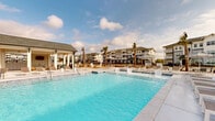 Located on Browning Way in Foley, Alabama, this outdoor amenity space delivers a resort-style experience designed for relaxation and recreation. The centerpiece is a generously sized swimming pool with crystal-clear blue water, featuring a gradual sun shelf entry that allows residents to lounge partially submerged in the shallow water. The pool deck is finished in a light-toned concrete surface and lined with an extensive collection of modern white-framed chaise lounges, providing ample space for sunbathing. Striped black-and-white market umbrellas are positioned throughout the deck to offer shaded retreats. A well-appointed single-story cabana structure anchors one side of the pool area, constructed with white board-and-batten siding, bold blue accent panels, and a pitched shingle roof. The covered porch of the cabana features ceiling fans and is furnished with additional lounge seating, offering a comfortable shaded gathering space. An outdoor bar or service area is also visible within the cabana pavilion. Tall palm trees are strategically placed throughout the space, reinforcing the coastal aesthetic and complementing the sunny Alabama Gulf Coast climate. A perimeter fence encloses the area, with multi-story residential buildings visible in the background, completing the community setting.