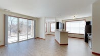 This single-floor space offers an open, airy layout that seamlessly connects the living and kitchen areas. Light hardwood flooring runs throughout, complementing the clean, neutral-toned walls and warm wood-trimmed windows and baseboards that add a touch of classic detail to the modern design. Large sliding glass doors on one side of the living area invite abundant natural light and provide views of the outdoor surroundings, while additional windows fitted with blinds further brighten the space. The kitchen features a central island with a built-in sink, offering both functional workspace and a natural gathering point. Overhead cabinetry in a dark finish provides a striking contrast to the otherwise light and neutral palette. A flush-mount ceiling light adds subtle illumination to complement the natural light. The open floor plan creates a generous sense of space, making the layout feel expansive and well-connected. The overall ambiance is clean, modern, and welcoming, with thoughtful design details that balance practicality with visual appeal throughout the entire floor.