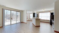 This single-floor space offers an open, airy layout that seamlessly connects the living and kitchen areas. Light hardwood flooring runs throughout, complementing the clean, neutral-toned walls and warm wood-trimmed windows and baseboards that add a touch of classic detail to the modern design. Large sliding glass doors on one side of the living area invite abundant natural light and provide views of the outdoor surroundings, while additional windows fitted with blinds further brighten the space. The kitchen features a central island with a built-in sink, offering both functional workspace and a natural gathering point. Overhead cabinetry in a dark finish provides a striking contrast to the otherwise light and neutral palette. A flush-mount ceiling light adds subtle illumination to complement the natural light. The open floor plan creates a generous sense of space, making the layout feel expansive and well-connected. The overall ambiance is clean, modern, and welcoming, with thoughtful design details that balance practicality with visual appeal throughout the entire floor.