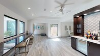 The WiFi Lounge is a thoughtfully designed communal workspace that blends modern aesthetics with functional comfort. The open-plan layout is anchored by clean white walls and light-toned porcelain tile flooring with subtle linear patterning, creating a bright and professional atmosphere throughout. A ceiling fan with brushed nickel finish is centrally positioned overhead, complementing recessed lighting that ensures consistent, well-distributed illumination across the space.

Along one wall, a sleek dark-toned counter with bar-style seating provides a casual workstation option, ideal for those seeking a more relaxed setup. On the opposite side, a private workstation with a dedicated desk, ergonomic chair, and dark cabinetry offers a more focused work environment. The dark wood cabinetry adds a warm contrast to the otherwise light and airy palette.

Natural light flows generously through large windows and a glass door that opens to an outdoor courtyard view, enhancing the sense of openness. A display wall showcasing colorful framed artwork adds a creative touch, while an adjacent alcove provides a transitional nook within the space. The overall ambiance is clean, contemporary, and conducive to both focused work and casual connectivity.