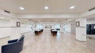 Located at 2401 6th St in Lake Charles, Louisiana, this single-floor community center offers a generously sized, open-plan interior designed to accommodate a variety of social and recreational activities. The main hall is expansive and bright, finished with light hardwood-style flooring and crisp white walls accented by a horizontal gray wainscoting band that runs throughout, lending the space a clean, cohesive aesthetic. Recessed ceiling lighting ensures the entire area is well-lit, complemented by natural light streaming in through multiple windows.

The main gathering area features several round tables and chairs arranged to support group activities and meetings, alongside a billiards table and a ping-pong table for recreational use. A large decorative artwork depicting a Louisiana heron and cypress landscape serves as a striking focal point on the main wall. A dedicated lounge nook offers comfortable navy leather seating arranged around a coffee table on a patterned area rug, ideal for informal conversations.

The kitchenette area is well-appointed with white cabinetry, granite countertops, a central island with bar-height seating, a black refrigerator, and a microwave — suitable for catered events or light food service. Adjacent to the kitchen, a resident mailbox bank and entry corridor provide practical building amenities, rounding out this fully functional and welcoming community space.